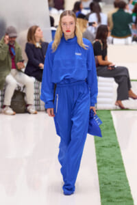 Female model in a bright blue Lacoste tracksuit walking down a fashion show runway with spectators in the background.