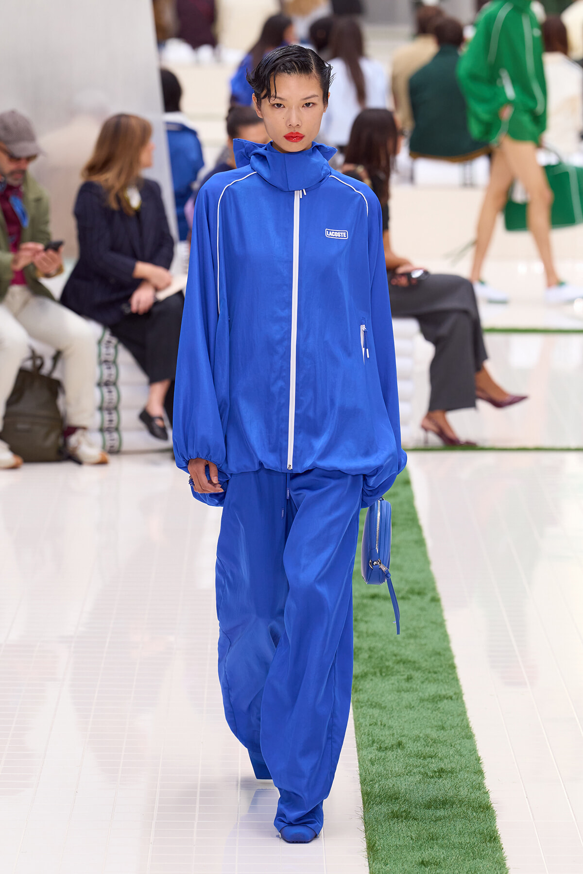 Runway model wearing a cobalt blue Lacoste tracksuit with white piping and a matching blue bag, walking a glossy white catwalk.