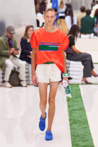 Model walks the runway in a bright orange T‑shirt, white shorts, and blue shoes, holding a white bag.
