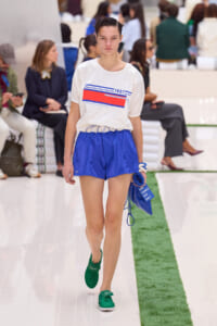 Model walking a fashion runway in a white graphic tee and blue shorts, carrying a blue pouch and wearing green sneakers.