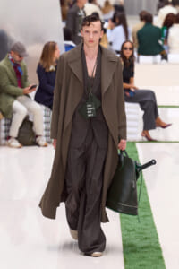 Male model on a runway wearing an olive oversized coat over a brown plaid suit, carrying a dark green bag with a 'FOR TENNIS USE ONLY' tag hanging from the neck