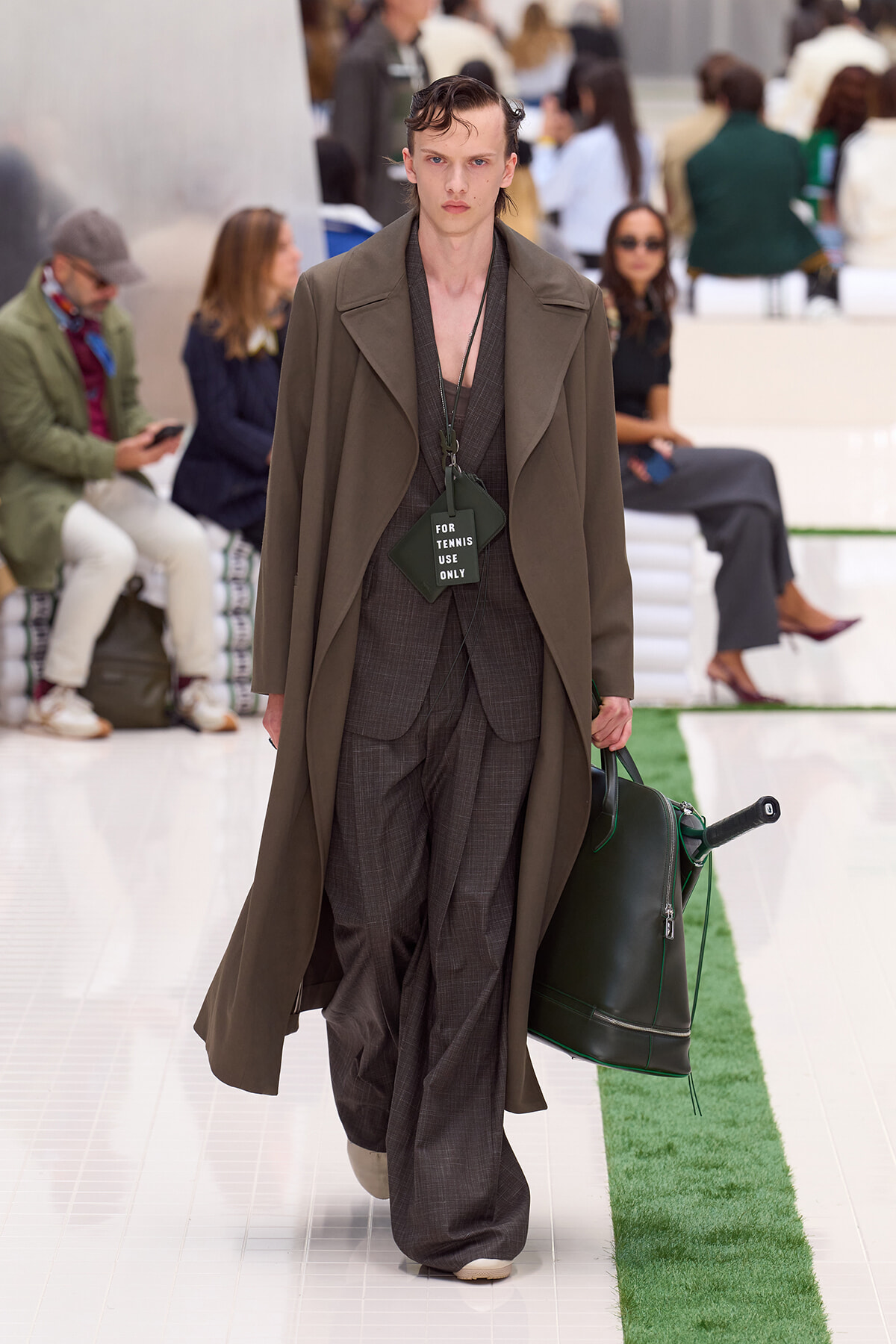 Male model on a runway wearing an olive oversized coat over a brown plaid suit, carrying a dark green bag with a 'FOR TENNIS USE ONLY' tag hanging from the neck