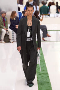 Male model walking the runway in a dark suit with a black tank, holding a white towel, backstage fashion show setting.