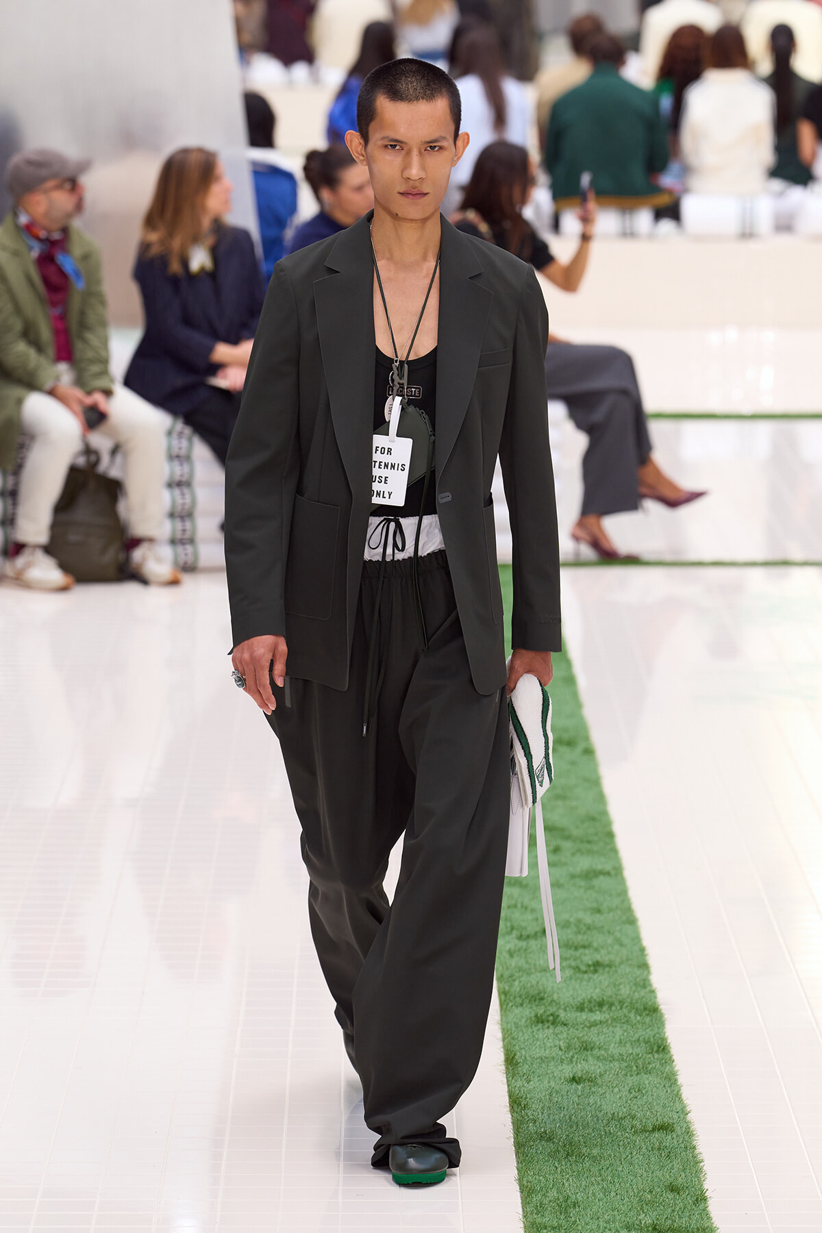 Male model walking the runway in a dark suit with a black tank, holding a white towel, backstage fashion show setting.