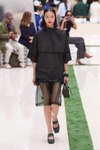 Model walking a fashion show in an oversized black top with a sheer green skirt beneath, carrying a small handbag and wearing dark green heels with striped socks đang (context: runway).