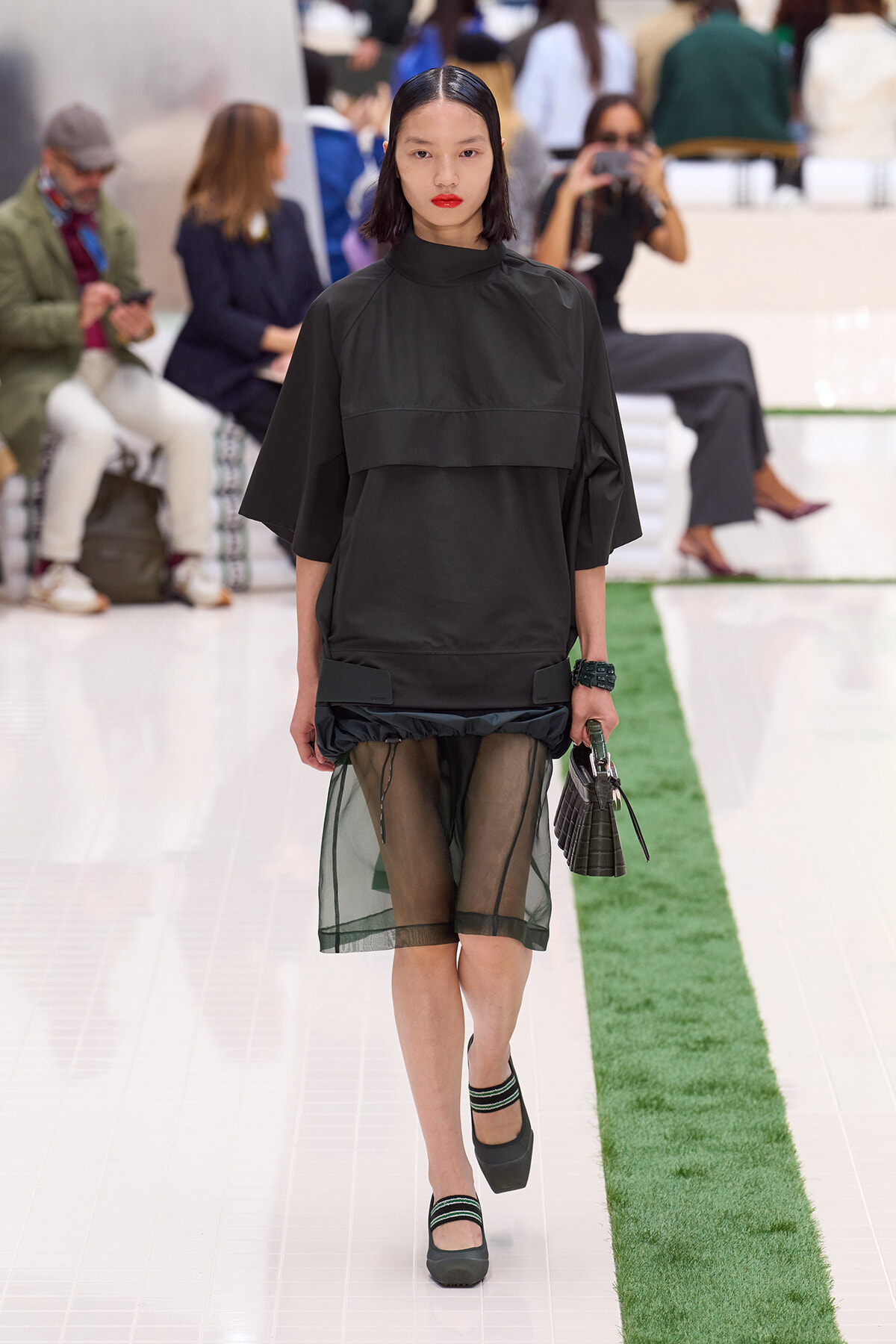 Model walking a fashion show in an oversized black top with a sheer green skirt beneath, carrying a small handbag and wearing dark green heels with striped socks đang (context: runway).