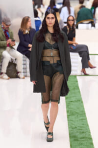 Model walks a runway in a sheer green dress under a dark coat, with layered necklaces and dark hair.
