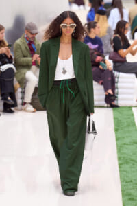 Model walking a runway in a forest-green blazer and loose matching pants with a white top and layered necklaces, wearing white sunglasses and carrying a white handbag.