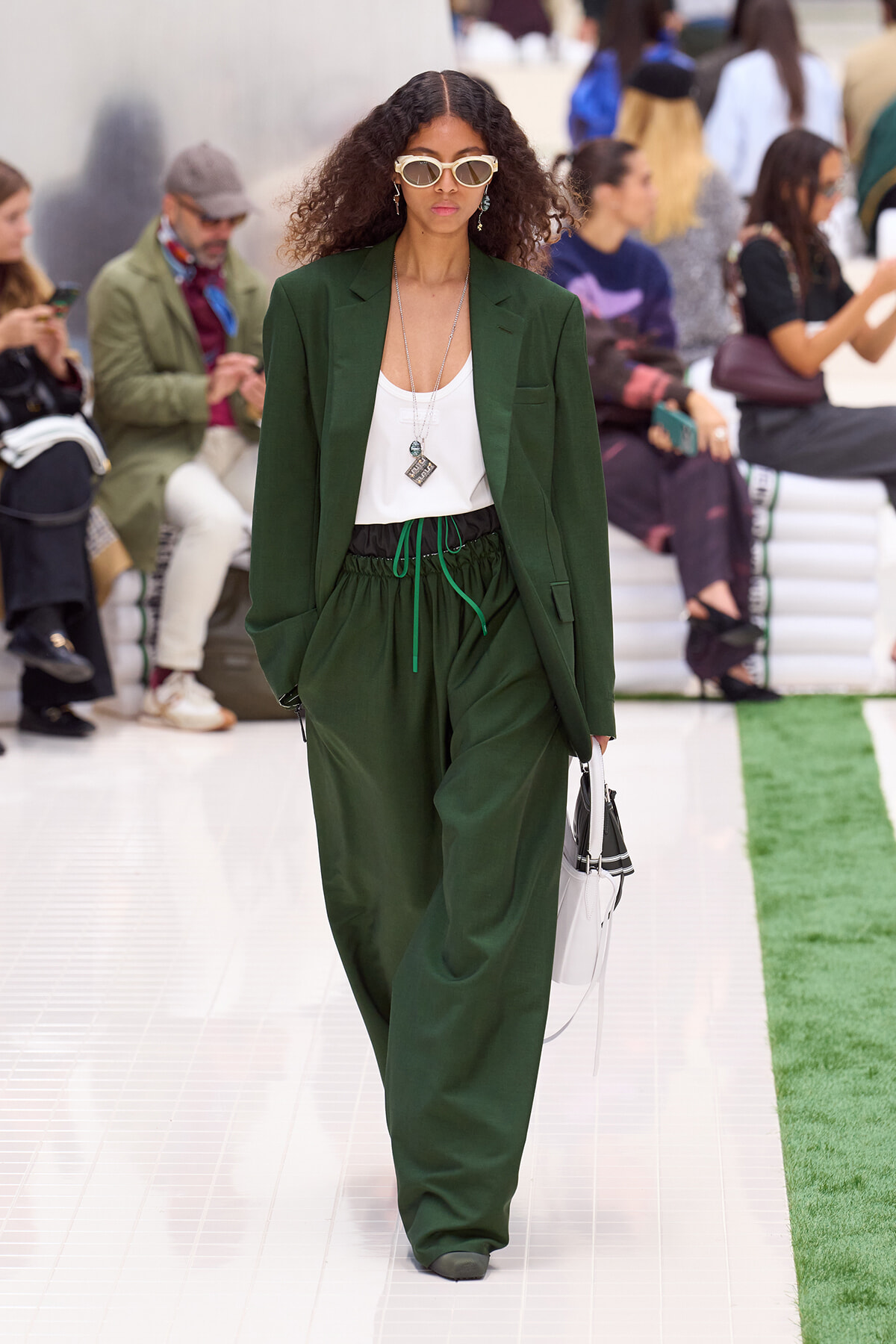 Model walking a runway in a forest-green blazer and loose matching pants with a white top and layered necklaces, wearing white sunglasses and carrying a white handbag.