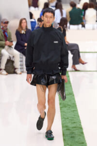 Male runway model in an all-black outfit (Lacoste top and glossy black shorts) walking the white catwalk with a green stripe in the foreground.