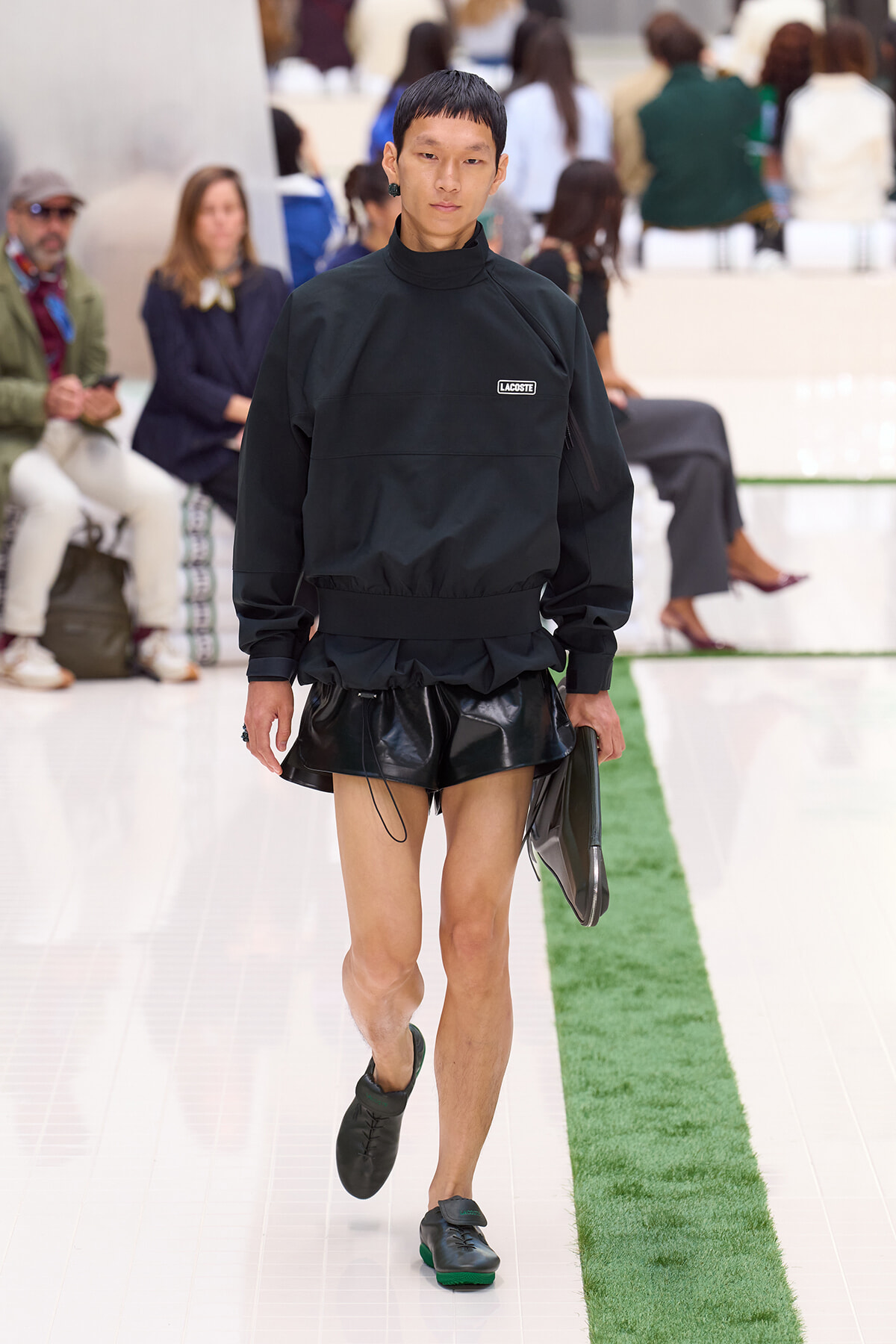 Male runway model in an all-black outfit (Lacoste top and glossy black shorts) walking the white catwalk with a green stripe in the foreground.