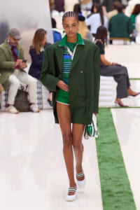Model walks a runway in a dark green oversized coat over a green striped top and green shorts, carrying a white bag with green accents.