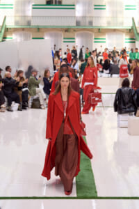 Model in a red coat and brown pants walks the fashion runway, spectators on both sides watching.