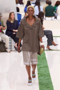 Model walking the runway in an oversized taupe top and beige textured shorts, holding a beige round clutch and wearing white sandals with green trim.
