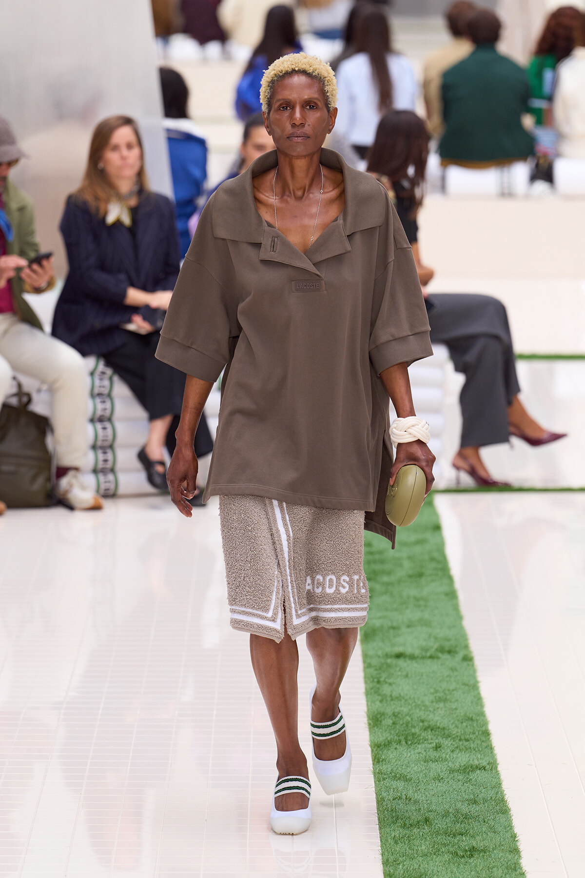 Model walking the runway in an oversized taupe top and beige textured shorts, holding a beige round clutch and wearing white sandals with green trim.