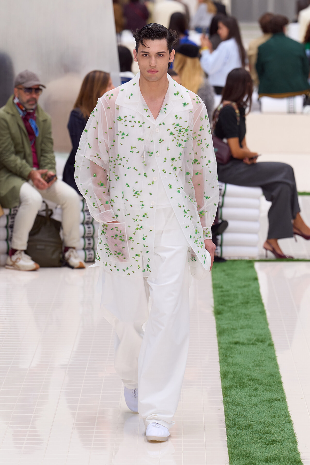 Male model walking a fashion runway in a sheer white shirt with green floral embroidery over white pants.