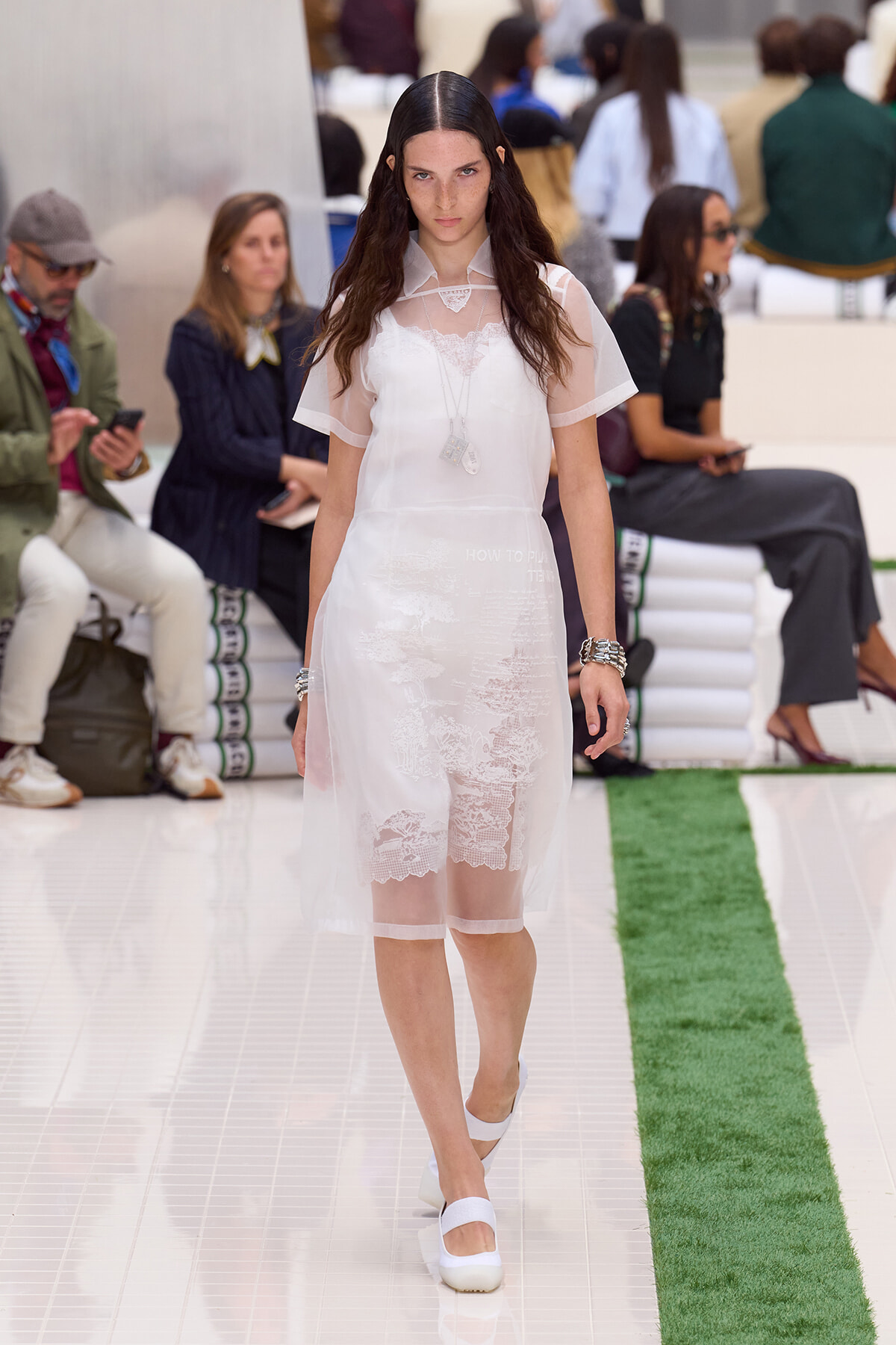 Young female model walks the white-tlo runway in a sheer white lace dress with a sheer cape, accessorized with layered necklaces and bracelets at a fashion show.