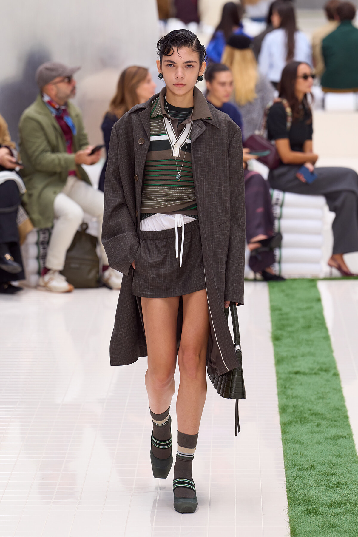 Model walks the runway in a brown plaid overcoat over a layered green striped top and brown shorts with a white drawstring, paired with striped socks and chunky heels, carrying a handbag.