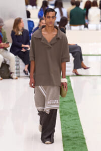 Male fashion model walks a runway in an oversized taupe polo with turned-down collar; he wears a textured beige towel-like wrap with 'ACOSTE' and dark wide pants, surrounded by seated audience.