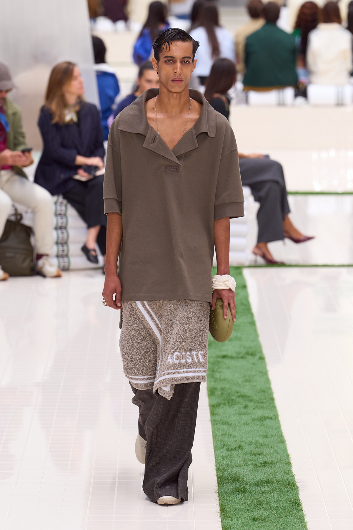 Male fashion model walks a runway in an oversized taupe polo with turned-down collar; he wears a textured beige towel-like wrap with 'ACOSTE' and dark wide pants, surrounded by seated audience.