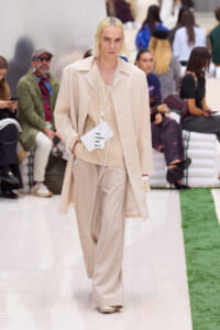 Fashion model walks a runway in a monochrome beige suit and oversized coat, with a white tag reading 'FOR TENNIS USE ONLY' hanging at the chest area.