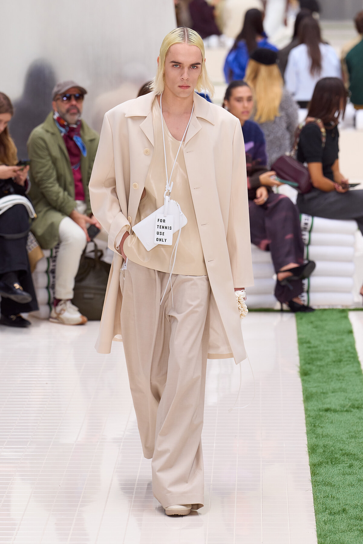 Fashion model walks a runway in a monochrome beige suit and oversized coat, with a white tag reading 'FOR TENNIS USE ONLY' hanging at the chest area.