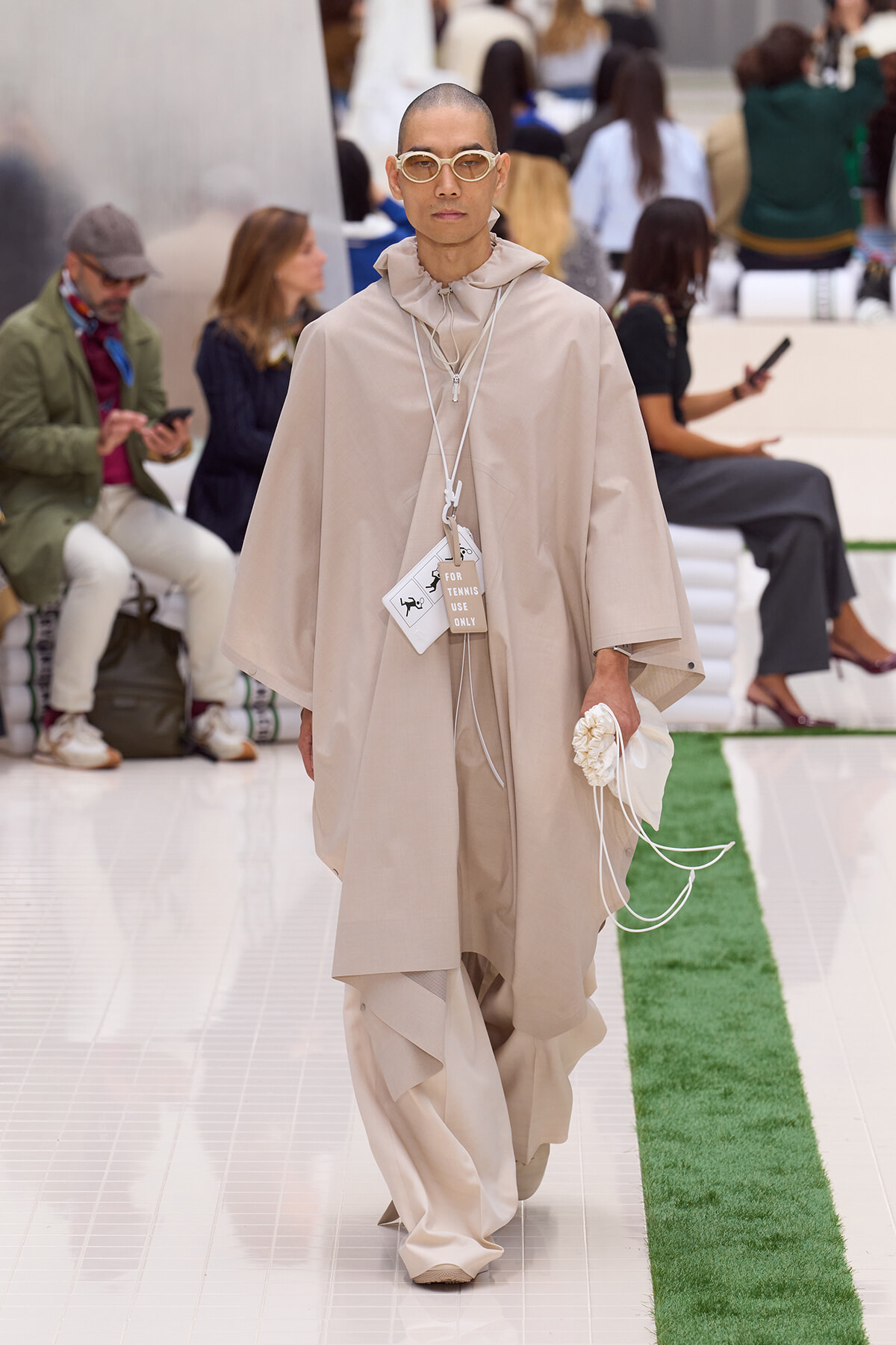 Model walking the runway in a loose beige cape and matching wide-leg pants, wearing rounded sunglasses with a tag hanging from the neck.