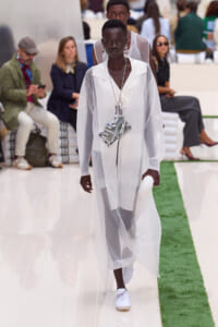 Model walks a sheer white ensemble on a runway, wearing layered silver necklaces and a metallic tag pendant, holding a white clutch as spectators watch.