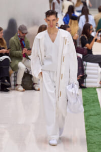 Male model on a white runway wearing an all-white ensemble with a long coat, V-neck top, and matching pants, carrying a white bag. Audience in background.