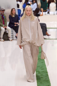 Model walks the runway in a beige oversized zip-front jacket and matching wide-leg pants, carrying a beige bag with a 'FOR TENNIS USE ONLY' tag.
