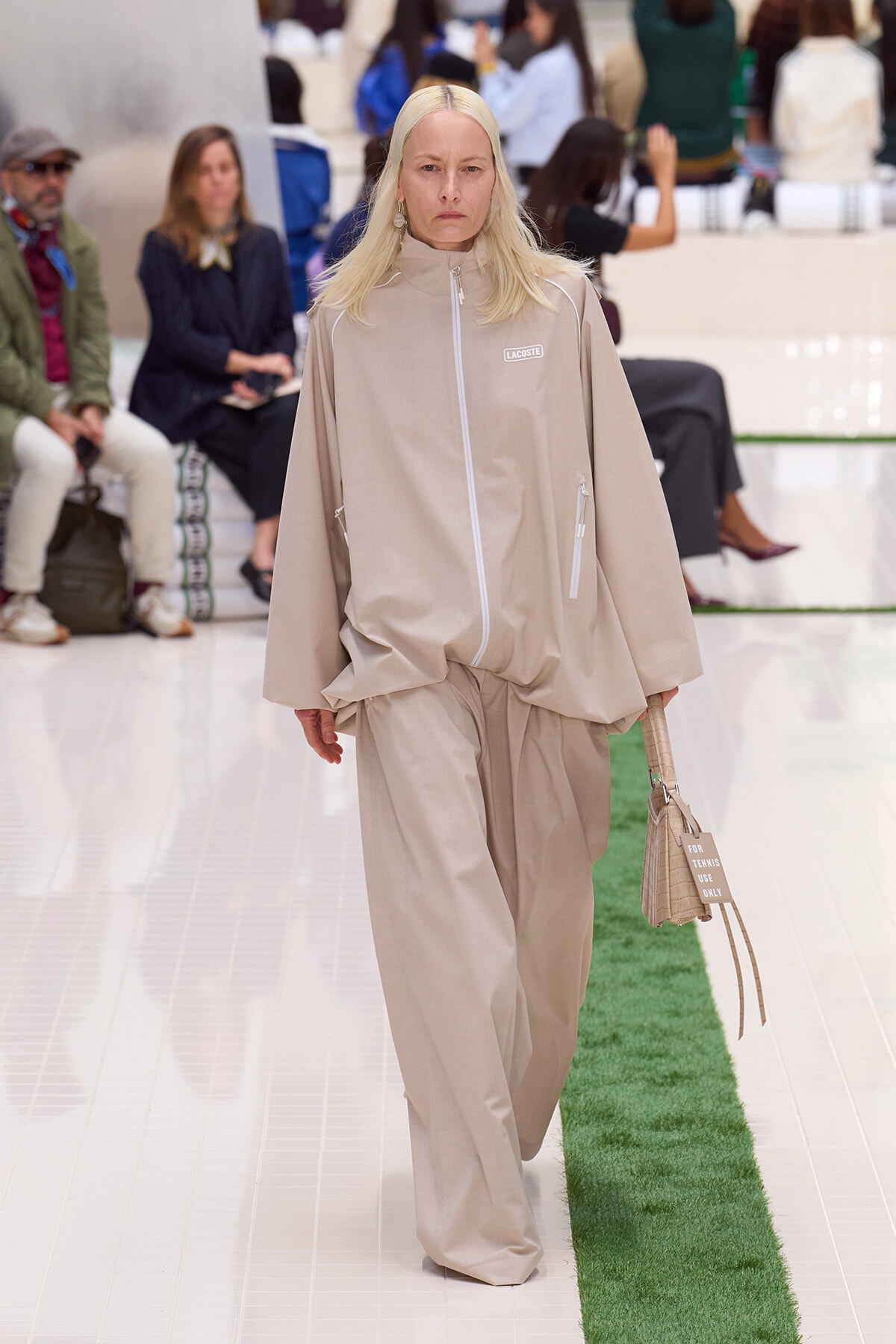 Model walks the runway in a beige oversized zip-front jacket and matching wide-leg pants, carrying a beige bag with a 'FOR TENNIS USE ONLY' tag.