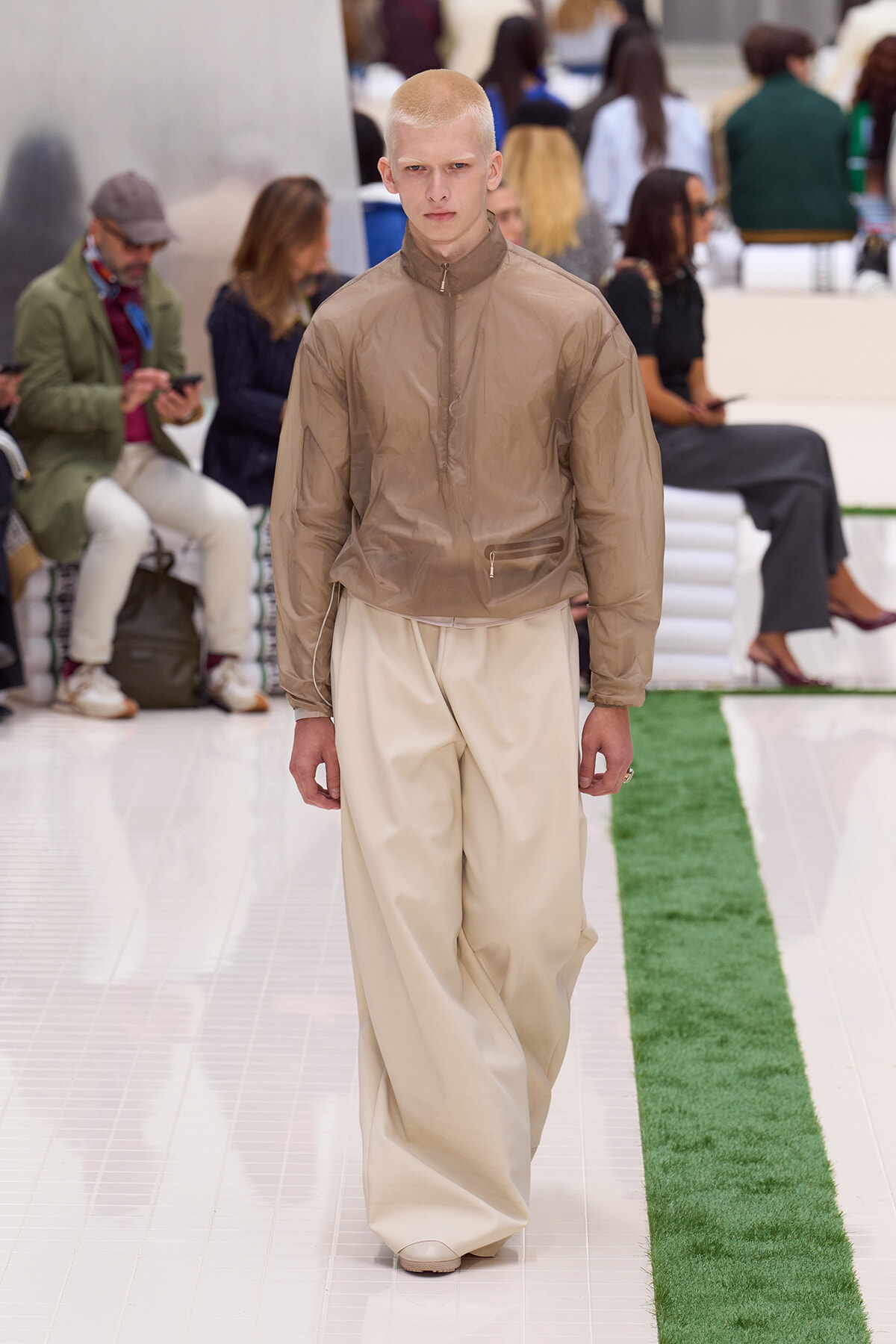 Model walking a runway in a taupe windbreaker and wide beige pants, with a focused expression.