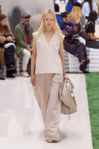 Model walks a beige minimalist outfit on a runway, carrying a large matching beige bag; seated audience visible in the background.