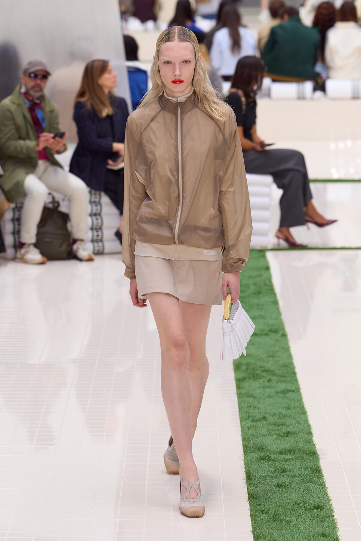 Model walks a runway in a beige windbreaker and matching mini skirt, carrying a small white handbag with a yellow handle.