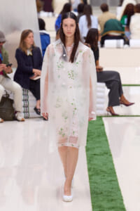 Female model walking a runway in a pale pink sheer coat with green floral embroidery, wearing a lace dress underneath.