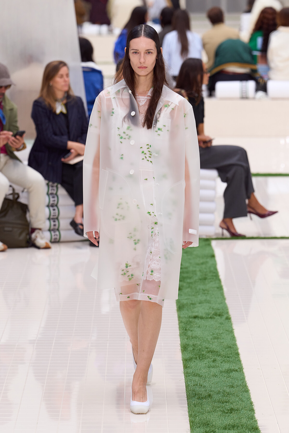 Female model walking a runway in a pale pink sheer coat with green floral embroidery, wearing a lace dress underneath.