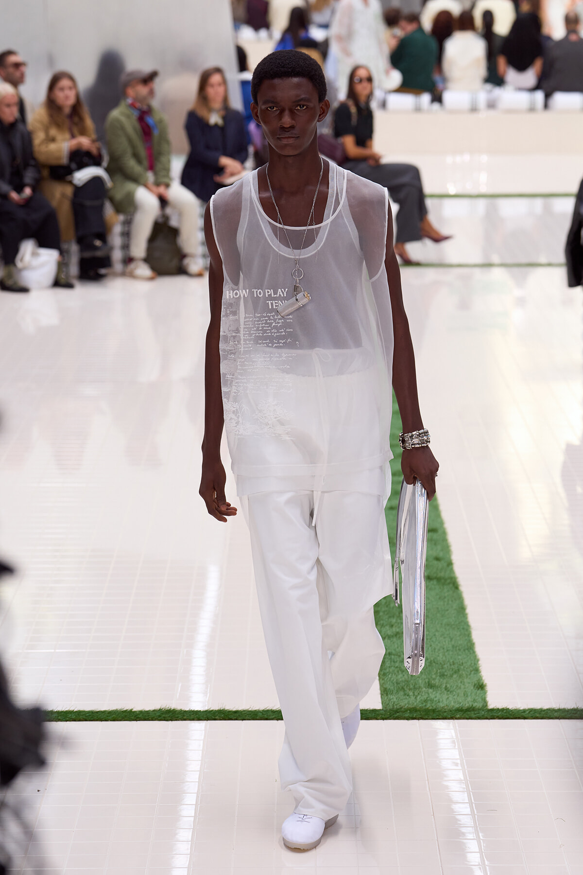 Male model walks a fashion runway in a sheer white sleeveless top and white wide pants, accessorized with silver jewelry against a fashion-show audience backdrop.