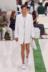 Male model walking a white double-breasted blazer and matching shorts down a glossy runway with a white clutch, dressed in white sneakers.