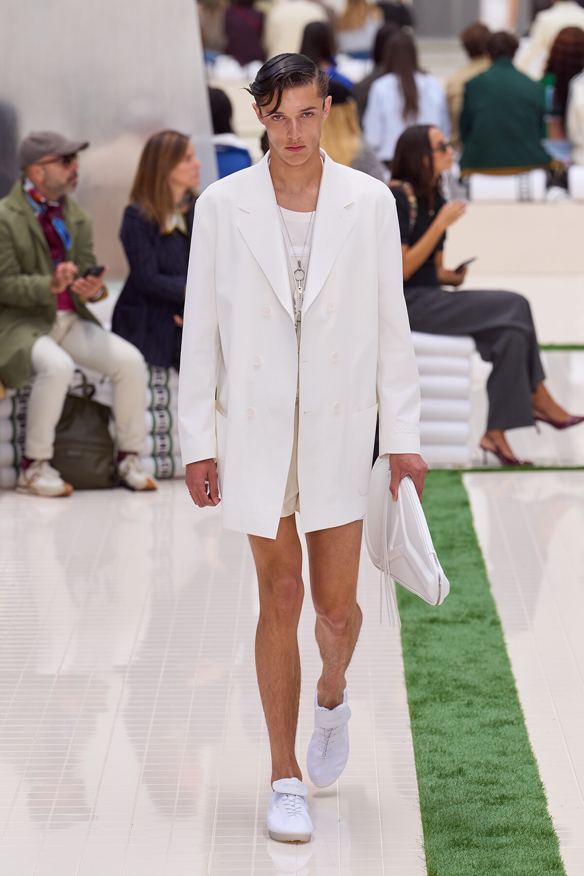 Male model walking a white double-breasted blazer and matching shorts down a glossy runway with a white clutch, dressed in white sneakers.