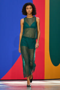 Model walking a runway in a green mesh sleeveless top and matching wide-leg pants, against a multicolor abstract backdrop.