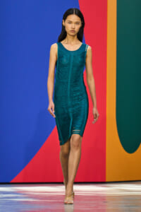 Model walking the runway in a teal textured sleeveless dress against a bold color-block backdrop on a fashion show catwalk