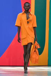 Model walking a runway in an orange shirt-dress with large pockets, carrying a matching orange bag, against a vivid color-block backdrop.