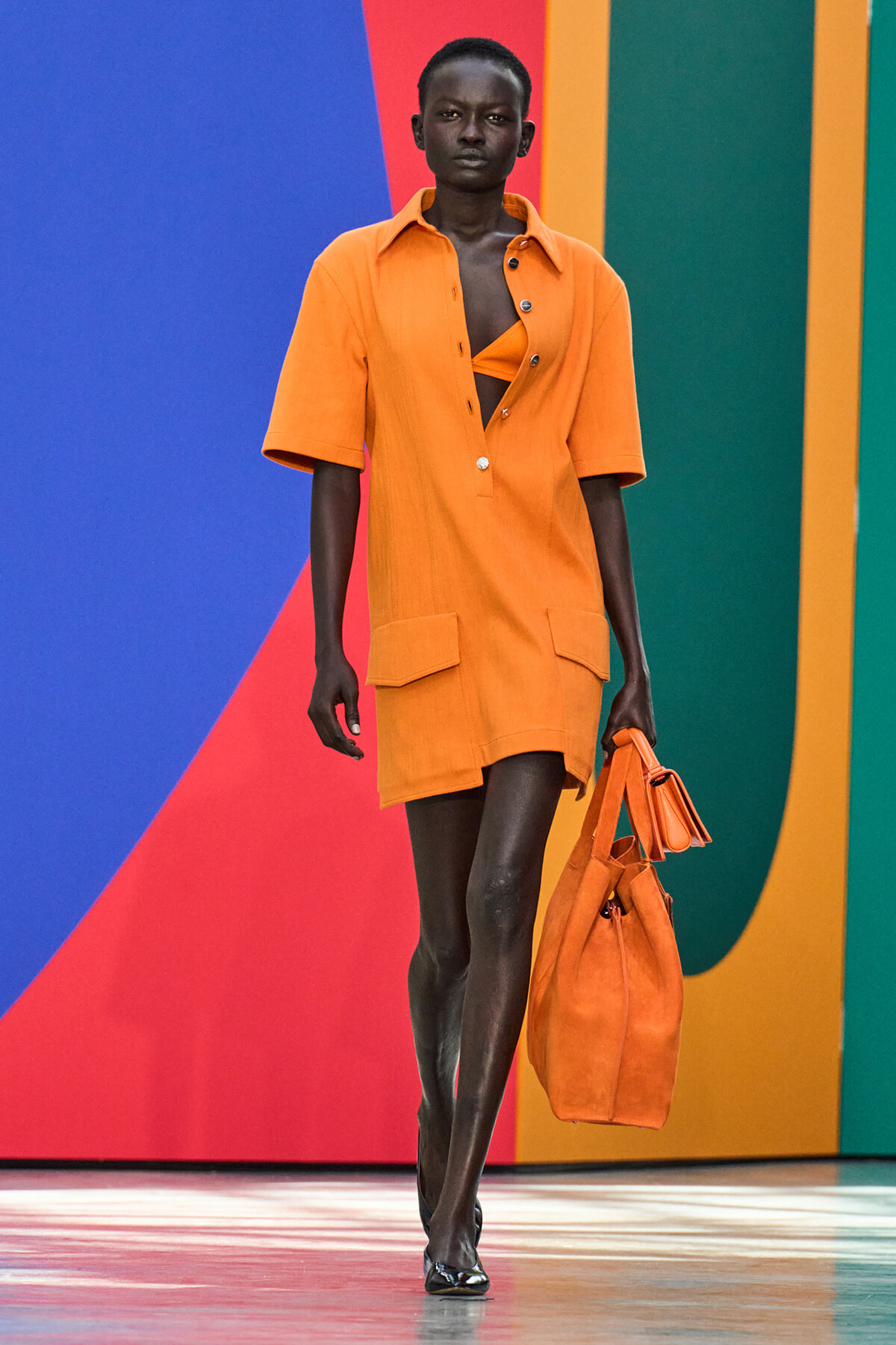 Model walking a runway in an orange shirt-dress with large pockets, carrying a matching orange bag, against a vivid color-block backdrop.