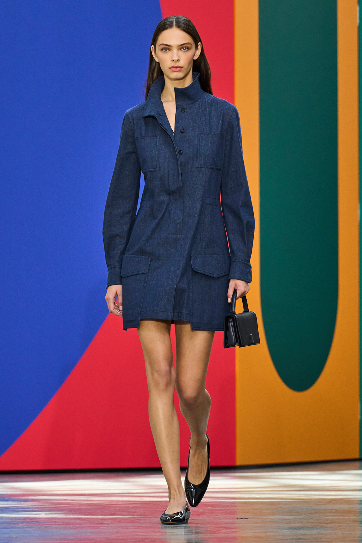Model walks the runway wearing a dark denim shirt-dress with a high collar and large front pockets, paired with black heels and a small black handbag against a bright, multicolor backdrop.
