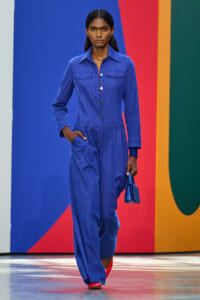 Female model walks a fashion runway wearing a cobalt blue denim jumpsuit with pockets, holding a small blue handbag, red shoes, against a bold color-block backdrop.