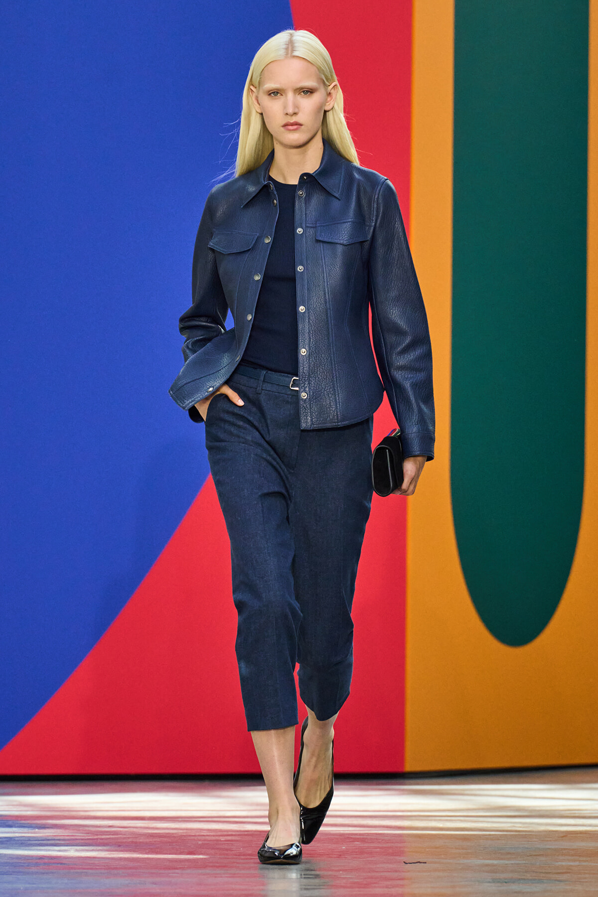 Female model walks a runway in a navy denim jacket and matching cropped jeans, holding a black clutch.