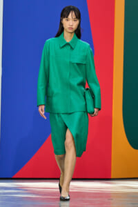 Model wearing a bright emerald green tailored suit with a knee-length skirt, walking on a multicolored runway backdrop.