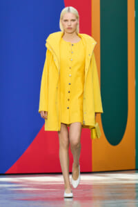 Model walks the runway in a bright yellow coat over a matching yellow dress, white heels, and a yellow clutch against a colorful geometric backdrop.