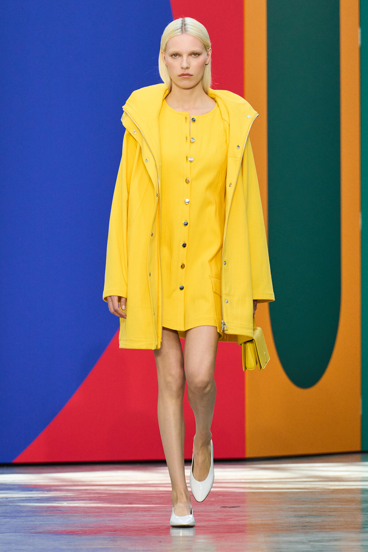 Model walks the runway in a bright yellow coat over a matching yellow dress, white heels, and a yellow clutch against a colorful geometric backdrop.