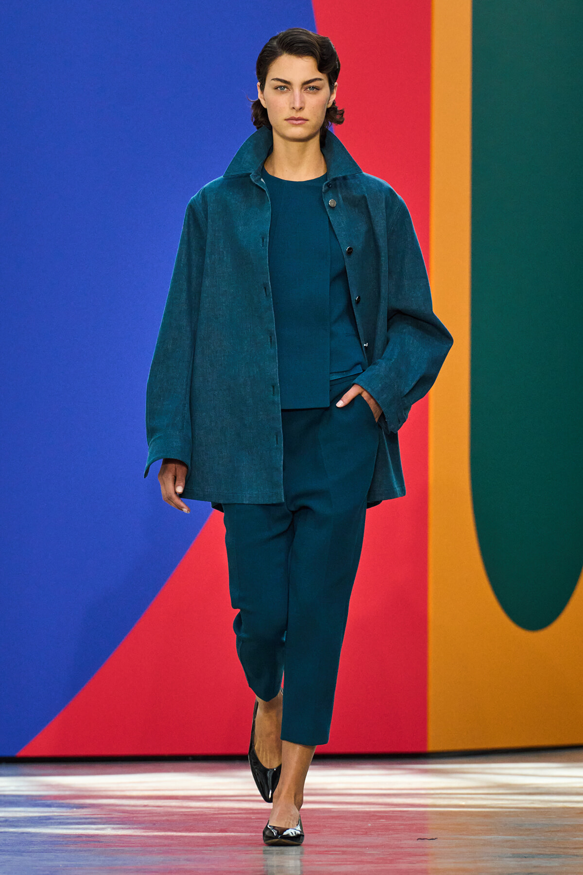 Model walking a teal outfit on a colorful runway background, wearing a loose teal coat and matching pants with black heels.